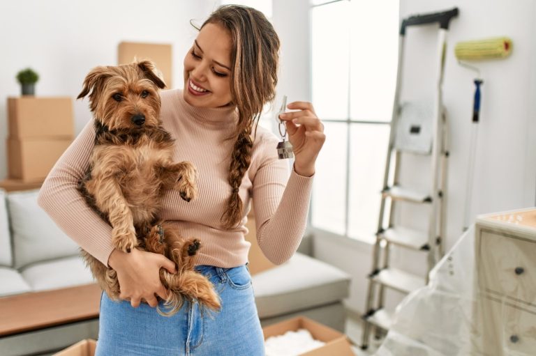 A woman holds a small dog, smiling, in a room with moving boxes and a ladder.