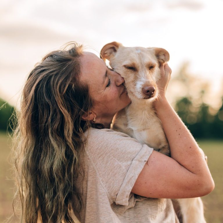 Woman with long hair hugging a light-coloured dog, smiling affectionately outdoors.